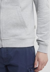 Hoodie gris zippé en tissu doux avec des poignets et une taille côtelés ; il possède une poche avant et une petite étiquette sur le côté.