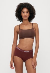 Brązowy biustonosz typu bralette z regulowanymi ramiączkami, w połączeniu z pasującymi szortami o wysokim stanie z białym logo "PIECES" na pasku.