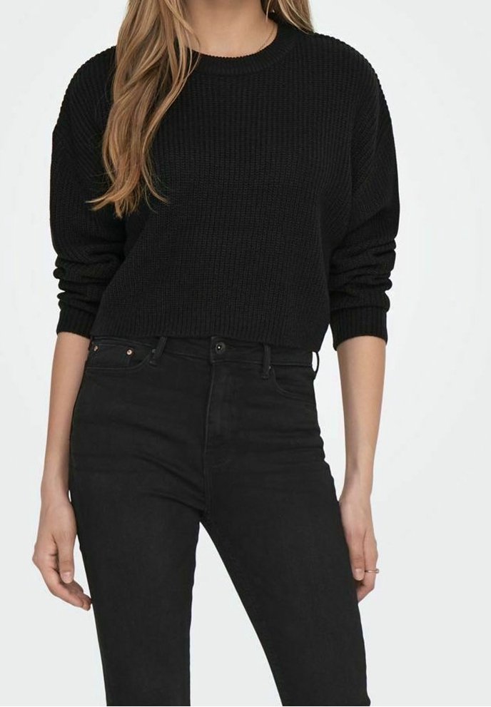 Femme portant un pull noir côtelé à manches longues et un jean noir taille haute, les bras détendus le long du corps.