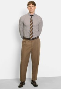 Herren-Anzug mit einem grau-weißen, leicht gestreiften Hemd, kombiniert mit braunen, gefalteten Hosen und dunklen Lederschuhen.