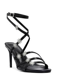 Sandales à talons hauts noires à lanières avec une finition brillante. Présente plusieurs ornements en forme de fleurs argentées le long des lanières et une fine boucle pour la cheville.