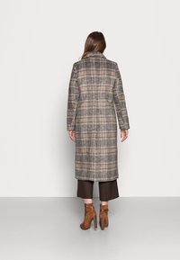 Šedý kostkovaný kabát s dlouhým, rovně střiženým designem, s klopovým límcem a bočními kapsami. V kombinaci s hnědými podpatkovými kotníkovými botami a kostkovanými kalhotami.