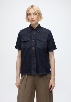 Personne aux cheveux blonds courts porte une chemise en denim foncé à manches courtes avec des boutons dorés et un pantalon marron à jambes larges.