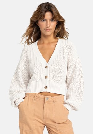 White knitted cropped cardigan with a V-neck, large buttons, and puffed sleeves, paired with beige pants.