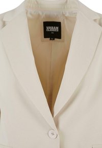 Blazer beige chiaro con colletto a revers, chiusura a un bottone e texture morbida; presenta un'etichetta interna con la scritta "URBAN CLASSICS" e taglia XS.