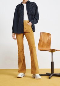 Schmal geschnittene Cord-Hosen in Senfgelb, kombiniert mit einem dunkelblauen Hemd und einem weißen, gerippten Top; im Hintergrund ist ein Holzstuhl zu sehen.