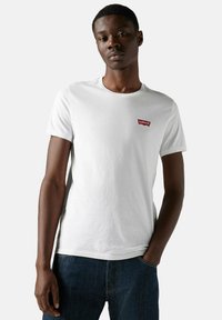 Ung mand iført en hvid Levi's t-shirt og mørke jeans, der står med den ene hånd i lommen imod en ensfarvet lys baggrund.