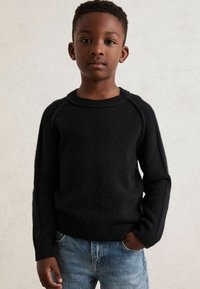 Schwarzer Strickpullover mit rundem Halsausschnitt und Raglanärmeln, kombiniert mit hellblauen Jeans. Die Textur ist weich und leicht fusselig.