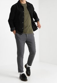 Muž ve černé bundě, olivově zeleném tričku, šedých kalhotách, bílých ponožkách a černých frněcích mokasínech stojící na bílém pozadí.