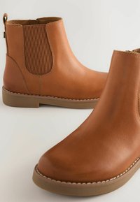 Une paire de bottines en cuir beige avec des panneaux élastiques sur les côtés et des semelles en caoutchouc brun sur un fond clair.