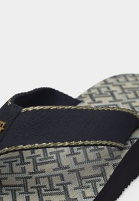 Κοντινή λήψη ενός σανδαλιού με μαύρο υφασμάτινο λουράκι που έχει χρυστή ραφή και σόλα με μοτίβο από διαδεμένες γεωμετρικές μορφές.