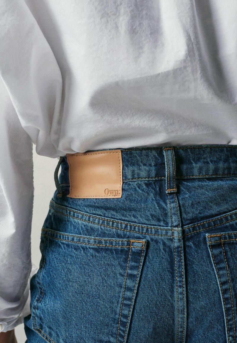 Personne portant un jean en denim bleu avec un patch en cuir beige à la ceinture et une chemise blanche ample à manches longues rentrée à l'arrière.