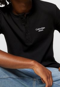Černé ribbené tričko henley se třemi knoflíky, na levé straně prsou s bílým logem "Calvin Klein Jeans".