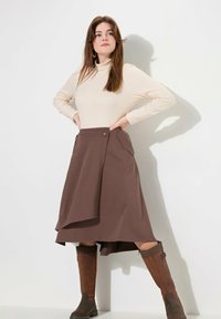 Brauner Wickelrock mit einem Knopfdetail, getragen über einem enganliegenden cremfarbenen Rollkragenpullover. Hohe braune Lederstiefel runden das Outfit ab.