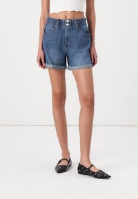 Kvinna klädd i uppvikta högmidjade blå denimshorts, vit croppad topp och svarta lägenheter med silveröglor, stående på ett vitt golv.