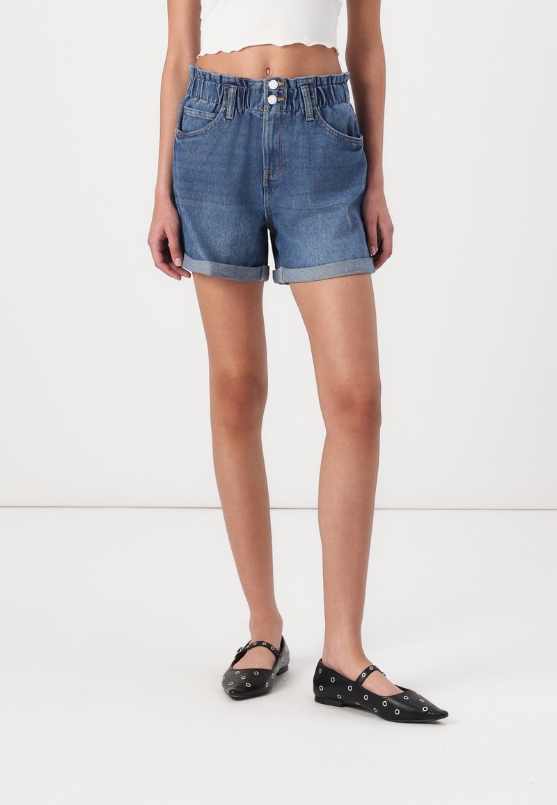 Kvinna klädd i uppvikta högmidjade blå denimshorts, vit croppad topp och svarta lägenheter med silveröglor, stående på ett vitt golv.