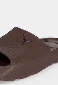 Sandale slide marron avec texture embossée et logo Jumpman noir sur la bride supérieure, conçue pour un usage décontracté.