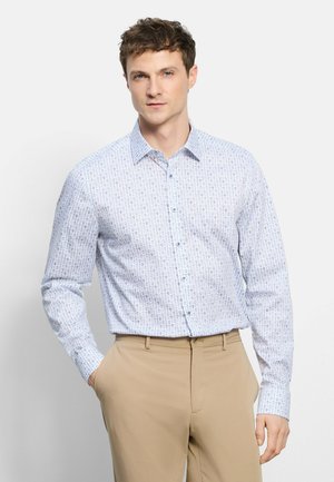 Homme portant une chemise à manches longues boutonnée bleu clair à motifs et un pantalon beige, une main dans la poche.