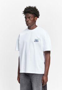 Weißes, oversized Baumwoll-T-Shirt mit rundem Ausschnitt, ausgestattet mit blauem Grafiktext und drei Figuren auf der linken Brustseite. Lässiger Schnitt, kurze Ärmel.