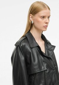 Juodas odinis švarkas su plačiu apykakle, sagstomu priekiu ir struktūruotais pečiais. Lygi tekstūra su subtiliu blizgesiu, pabrėžianti pritaikytą dizainą.