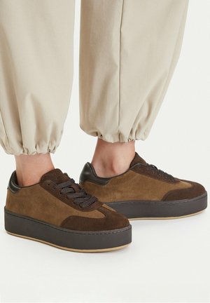 Zapatillas de plataforma de ante marrón con cordones negros, usadas con pantalones beige con puños, mostradas de lado sobre un fondo claro.
