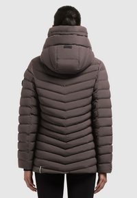Donna che indossa un giubbotto imbottito color taupe scuro con un grande cappuccio, trapuntato a zig-zag, e leggins neri, vista da dietro su uno sfondo bianco.