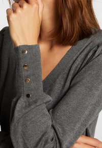 Pull gris en maille avec un col en V, poignets côtelés ornés de quatre boutons dorés. Texture douce et design décontracté avec des manches légèrement bouffantes.