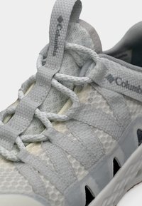 Αθλητικό παπούτσι από ανοιχτό γκρι δίχτυ με υφασμάτινες επιφάνειες, ενσωματωμένο σύστημα κορδονιών, σήμανση Columbia και κομμένα τμήματα στη σόλα για αερισμό.