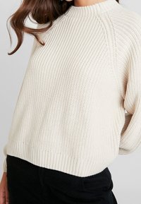 Pull en maille couleur crème avec texture côtelée, col montant et coutures d'épaules tombantes. Doté d'une taille ajustée pour une coupe décontractée.