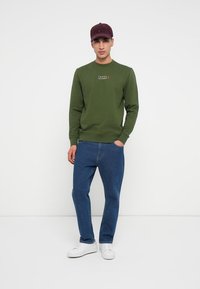 Muž v bordovom klobúku, olivovozelenom mikinovom svetri Tommy Hilfiger, modrých džínoch a bielych teniskách stojí pred bielym pozadím.