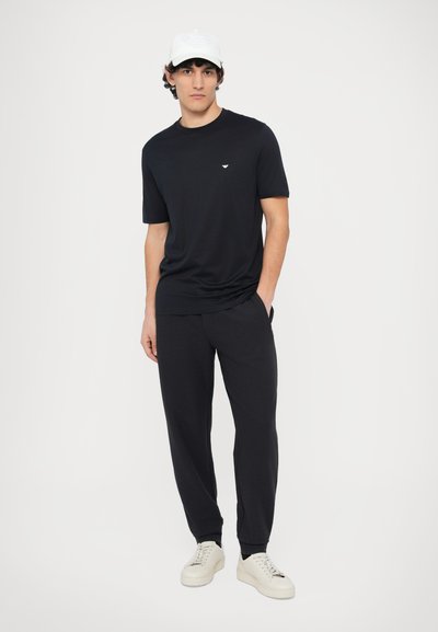 Čierne tričko s krátkym rukávom s malým logom, v kombinácii s textúrovanými čiernymi nohavicami a bielymi teniskami. Jednoduchý a moderný dizajn.