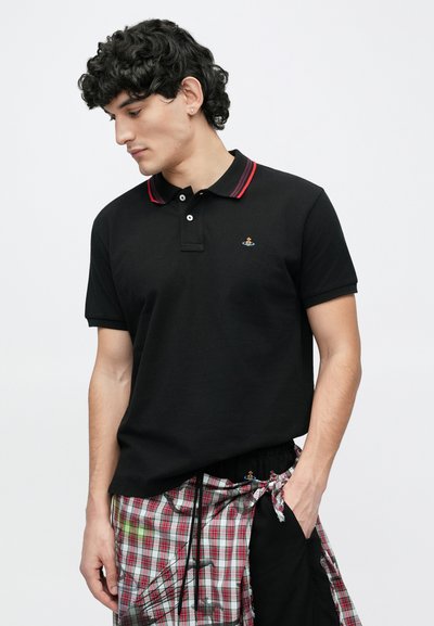 Moški s kodrastimi črnimi lasmi, oblečen v črno polo majico z rdečim robom in kariraste hlače, gleda navzdol z eno roko v žepu na belem ozadju.