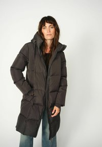 Lang pufferjakke, mørkegrå, med hætte. Har lynlås foran, store påsatte lommer og et quiltet design. Blød, polstret tekstur.