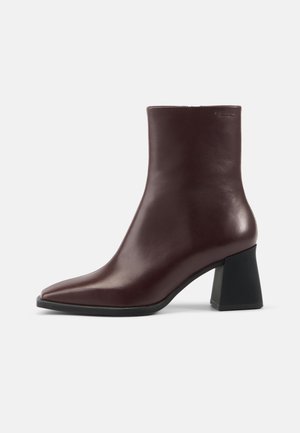 Dunkelbrauner Lederknöchelhoher Stiefel mit spitzer Spitze und schwarzem Blockabsatz auf schlichtem hellem Hintergrund.