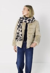 Vrouw met beige nep bontjas, zwarte top, blauwe spijkerbroek en een zwart-witte gevlekte sjaal, kijkt opzij tegen een witte achtergrond.