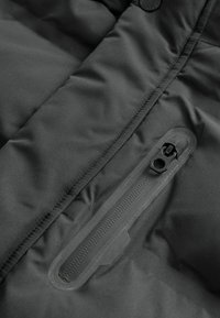 Gros plan sur une poche de veste imperméable noire avec une fermeture éclair scellée et un détail en tissu matelassé cousu.