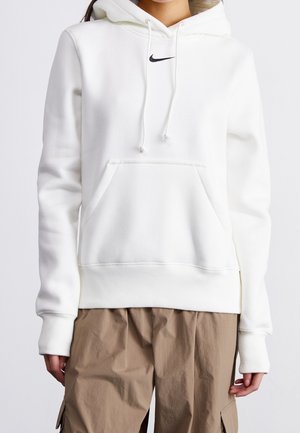 Persoon draagt een witte Nike hoodie met voorzak en trekkoorden, gecombineerd met loszittende lichtbruine cargobroek.