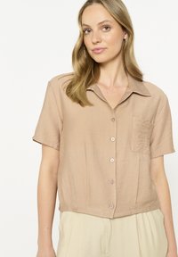 Short-sleeve, collared shirt in beige fabric with a textured finish, featuring a pocket and button closure along the front.