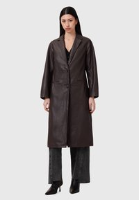 Cappotto lungo in pelle marrone con colletto a doppio abbottonatura, due bottoni e tasche laterali, abbinato a pantaloni larghi grigi e tacchi a punta neri.