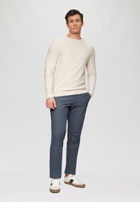 Beige gestrickter Pullover, blaue gemusterte Hose mit feiner Textur und weiße Sneaker mit dunklen Akzenten. Lässiges Outfit an einem stehenden Modell.