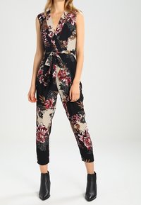 Blommig jumpsuit med en svart och beige färgkombination, omslagdesign, ärmlös topp och knytning i midjan, i kombination med svarta ankboots.
