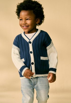 Marineblauer und weißer gerippter Cardigan mit Knopfleiste, zwei Fronttaschen und kontrastierenden Ärmeln. Kind trägt ihn mit hellen Jeans.
