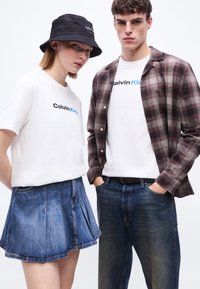 Δύο νέοι φορώντας λευκά t-shirts Calvin Klein. Ο ένας έχει πουκάμισο σε καρό μοτίβο και τζιν, ο άλλος φοράει μαύρο καπέλο τύπου bucket και τζιν φούστα.