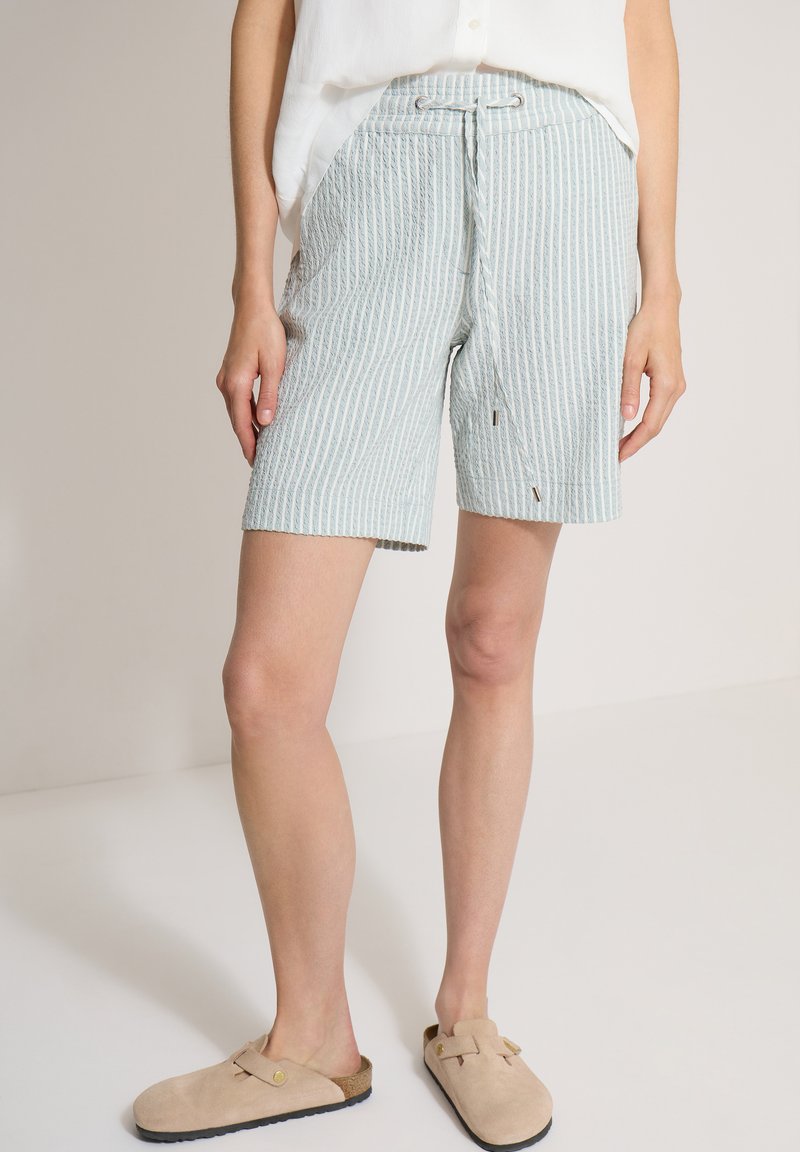 Personne portant un short à rayures verticales bleu clair et blanc avec cordon de serrage et chaussures beige à enfiler sur un fond uni.