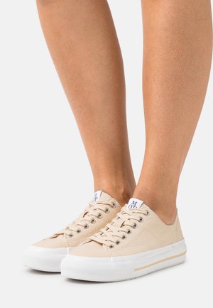Beige canvas sneakers with white soles, round toe, and lace-up design. Features understated logo on the tongue and side. Smooth texture.