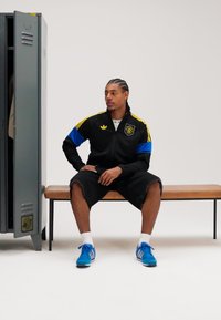 Melnas Adidas sporta jaka ar dzeltens un zils akcentiem, melnas šorti un zili sporta apavi. Sēžot uz koka sola pie pelēka skapīša.