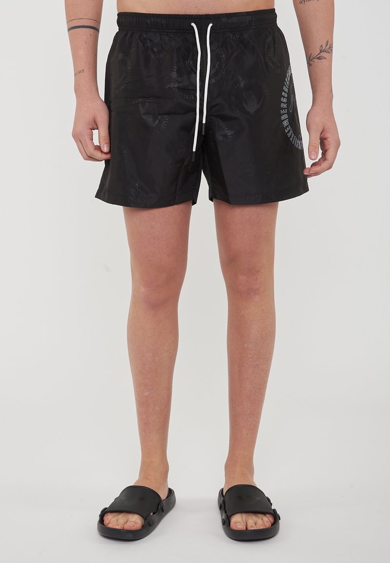 Costumi da bagno neri con elastico in vita, cordoncino bianco e una sottile texture a motivo. Indossati con sandali neri a infradito.