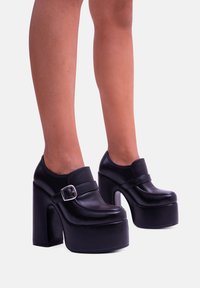 Scarpe platform nere in pelle liscia, con un ampio tacco a blocco, punta arrotondata e un dettaglio con fibbia sulla cinghia anteriore.
