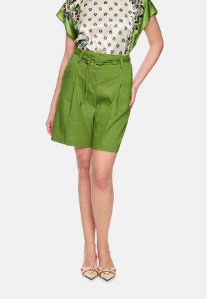 Frau trägt grüne High-Waist-Shorts mit Gürtel, blumige gemusterte Seidenbluse und spitze gewebte Absätze mit Schnallen, steht vor weißem Hintergrund.