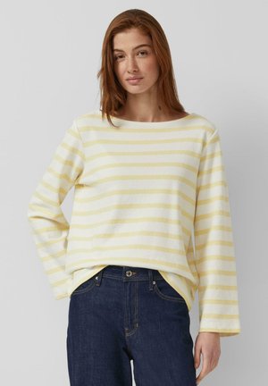 Femme aux cheveux roux portant un t-shirt à manches longues rayé jaune et blanc et un jean bleu foncé, debout devant un fond gris uni.